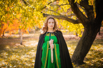 A beautiful girl in a medieval green dress with gold braid holds a dagger in her hands. Queen in a cloak and in a blio dress in the autumn forest.