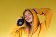 © Igor - Funny young girl looking, waving hand, holding portable wireless bluetooth music speaker, isolated on yellow background. Girl in a yellow hoodie