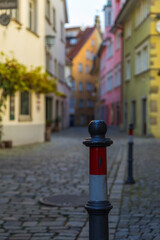 Im Vordergrund befindet sich ein Strassenbegrenzungspfosten an einer gepflasterten Strasse und im unscharfen Hintergrund eine bunte Häuserfront.