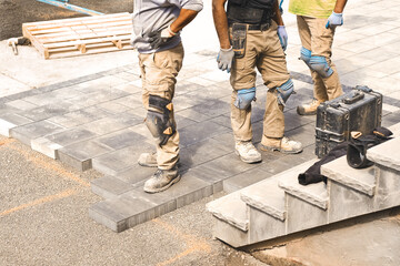 Three construction workers working on landscaping construction site, laying paving interlock stones slabs for quality patio stonework, home renovation project. Prospective view of man onsite job.