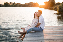 A Husband With A Pregnant Wife Is Resting On The River Bank, Sitting On A Pier In Summer Sunny Weather At Sunset. Couple In Love Man And Woman Hugging, Holding Hands,