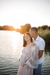 © DSMT - A pregnant woman and her husband stand on the pier and look at each other, a beautiful river and sunset, future parents spend time outdoors, a beautiful young mother