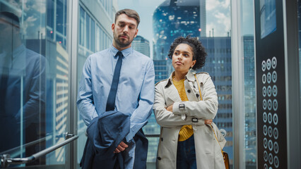 Wall Mural - Two Office Colleagues Riding in Glass Elevator in a Modern Business Center on the Way From Work. Caucasian Male Specialist and Black Latin American Female Manager Casually Chat in the Lift.