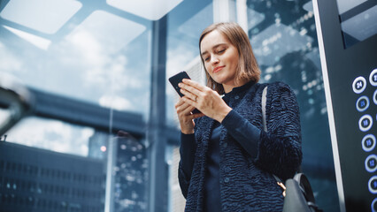 Wall Mural - Confident Young Adult Female Riding Glass Elevator to Office in Modern Business Center. Successful Manager Using Smartphone, Write Message, Check Social Media and Emails in a Lift.