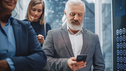 Wall Mural - Successful Middle Aged Businessman Riding Crowded Glass Elevator to Office in Modern Business Center. Handsome Happy Man Using Smartphone, Write a Message, Social Media and Work Emails in a Lift.