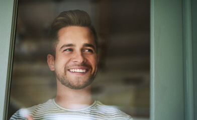 Wall Mural - Make everyday an awesome one. Shot of a handsome young man looking out of a window at home and smiling.