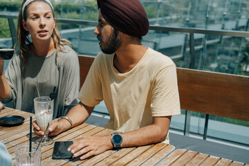 Two Multi Ethnic Friends Drinking Coffee in the Outdoor Cafe. 
Serious young woman holding cup of espresso and talking to Indian man with turban and iced coffee while sitting together at table outside