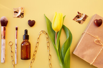 Beauty card concept. Cosmetics, accessories on pastel yellow background with chocolate hearts and yellow tulip. Trendy glasses, makeup brushes, serum dropper, chain, earrings and gift box.