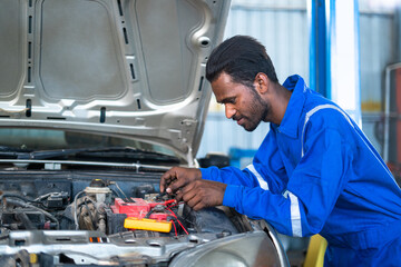 Car mechanic testing or checking car engine battery by using digital multimeter at garage - concept of technology, maintenance services and blue collar jobs.