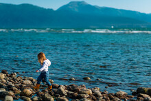 A Young Girl In Rain Boots Walks Along The Rocky Shore In Front Of A Mountain In Alaska
