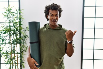 Poster - African man with curly hair holding yoga mat at studio surprised pointing with hand finger to the side, open mouth amazed expression.