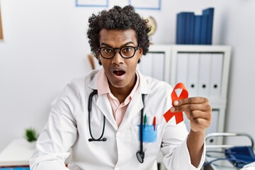 Sticker - African doctor man holding support red ribbon scared and amazed with open mouth for surprise, disbelief face