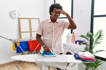 Poster - African man with curly hair ironing clothes at home very happy and smiling looking far away with hand over head. searching concept.