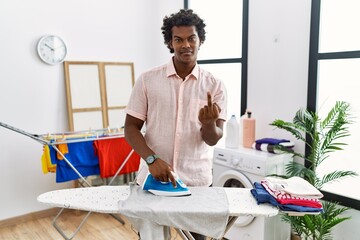 Poster - African man with curly hair ironing clothes at home showing middle finger, impolite and rude fuck off expression