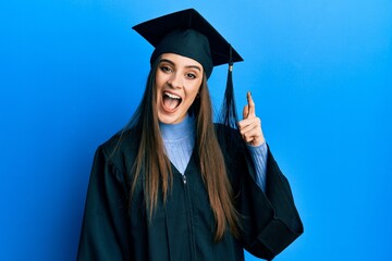 Wall Mural - Beautiful brunette young woman wearing graduation cap and ceremony robe pointing finger up with successful idea. exited and happy. number one.
