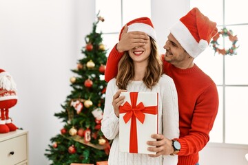 Wall Mural - Young hispanic couple wearing christmas hat. Man surprising his girlfriend with hands on eyes and gift at home.