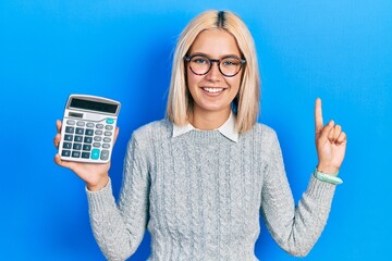 Sticker - Beautiful blonde woman showing calculator device smiling with an idea or question pointing finger with happy face, number one