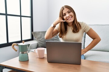 Canvas Print - Young brunette woman using laptop at home drinking a cup of coffee smiling doing phone gesture with hand and fingers like talking on the telephone. communicating concepts.