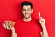 © Krakenimages.com - Young hispanic man holding peanuts smiling with an idea or question pointing finger with happy face, number one