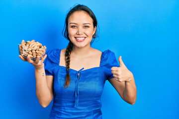 Wall Mural - Young hispanic girl holding bowl of peanuts smiling happy and positive, thumb up doing excellent and approval sign