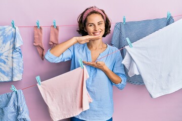 Sticker - Young brunette woman doing laundry around string hangs gesturing with hands showing big and large size sign, measure symbol. smiling looking at the camera. measuring concept.