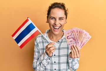 Poster - Young brunette woman holding thailand flag and baht banknotes smiling and laughing hard out loud because funny crazy joke.