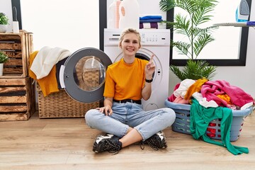 Poster - Young blonde woman doing laundry sitting by washing machine showing and pointing up with finger number one while smiling confident and happy.