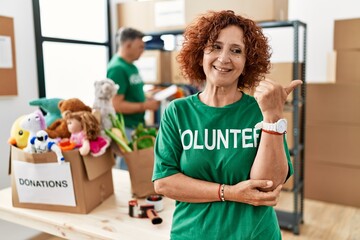 Poster - Middle age woman wearing volunteer t shirt at donations stand smiling with happy face looking and pointing to the side with thumb up.