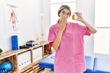 Wall Mural - Young physiotherapist woman working at pain recovery clinic holding pills covering mouth with hand, shocked and afraid for mistake. surprised expression