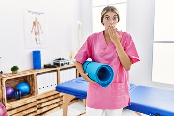 Wall Mural - Young physiotherapist woman working at pain recovery clinic holding yoga mat covering mouth with hand, shocked and afraid for mistake. surprised expression