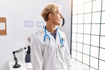 Poster - Young blond man wearing doctor uniform and stethoscope at clinic looking to side, relax profile pose with natural face and confident smile.