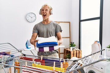 Poster - Young blond man listening to music doing laundry angry and mad screaming frustrated and furious, shouting with anger. rage and aggressive concept.