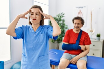 Poster - Middle age doctor woman with patient with arm injury at rehabilitation clinic with hand on head, headache because stress. suffering migraine.