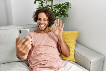 Sticker - Young hispanic man smiling happy having video call using smartphone sitting on the sofa at home.