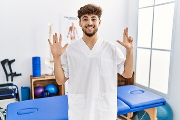 Wall Mural - Young arab man working at pain recovery clinic showing and pointing up with fingers number eight while smiling confident and happy.