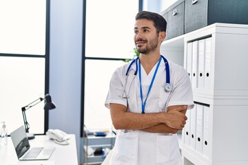 Wall Mural - Young hispanic man wearing doctor uniform standing with arms crossed gesture at clinic