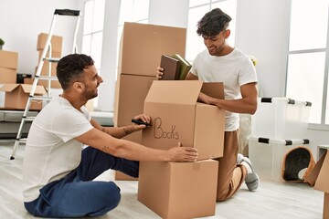 Two hispanic men couple smiling confident packing book cardboard box at new home