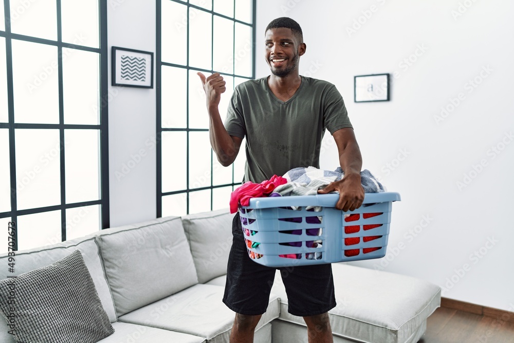 Young african american man doing laundry holding basket pointing thumb up to the side smiling happy with open mouth