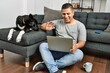 © Krakenimages.com - Young hispanic man having video call using laptop sitting on the floor with dog at home.