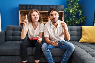 Canvas Print - Young couple playing video game holding controller at home surprised with an idea or question pointing finger with happy face, number one