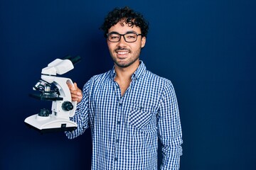 Poster - Young hispanic man holding microscope looking positive and happy standing and smiling with a confident smile showing teeth