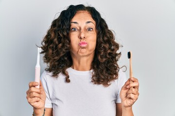 Canvas Print - Middle age hispanic woman holding electric toothbrush and teethbrush puffing cheeks with funny face. mouth inflated with air, catching air.