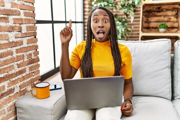 Poster - African woman sitting on the sofa using laptop at home pointing finger up with successful idea. exited and happy. number one.