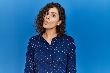 Poster - Young brunette woman with curly hair wearing casual clothes over blue background making fish face with lips, crazy and comical gesture. funny expression.