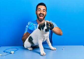 Wall Mural - Handsome hispanic veterinary man with beard putting vaccine to puppy dog smiling and laughing hard out loud because funny crazy joke.