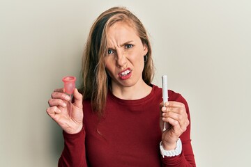 Young blonde woman holding menstrual cup and tampon in shock face, looking skeptical and sarcastic, surprised with open mouth