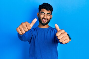 Wall Mural - Arab man with beard wearing casual blue sweater approving doing positive gesture with hand, thumbs up smiling and happy for success. winner gesture.