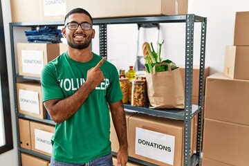 Wall Mural - Young indian man volunteer holding donations box cheerful with a smile of face pointing with hand and finger up to the side with happy and natural expression on face