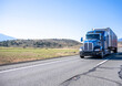 Dark blue big rig semi truck with extended cab with roof windows transporting cargo in covered framed semi trailer running on the highway road with hills on the side in California