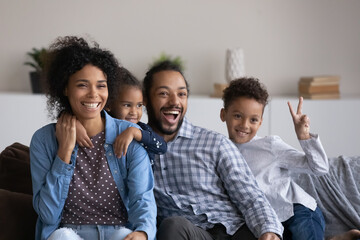 Wall Mural - Excited joyful Black family with two cute sibling boy and girl having fun together at home, resting on couch, laughing, shouting, showing victory. African parents and little kids portrait